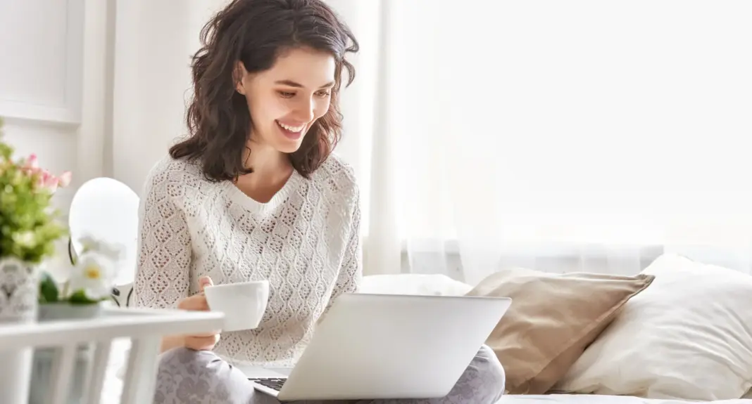 Wanita bahagia duduk di tempat tidur dengan kopi dan laptop, simbol pengguna Windows 10 yang perlu mempertimbangkan upgrade