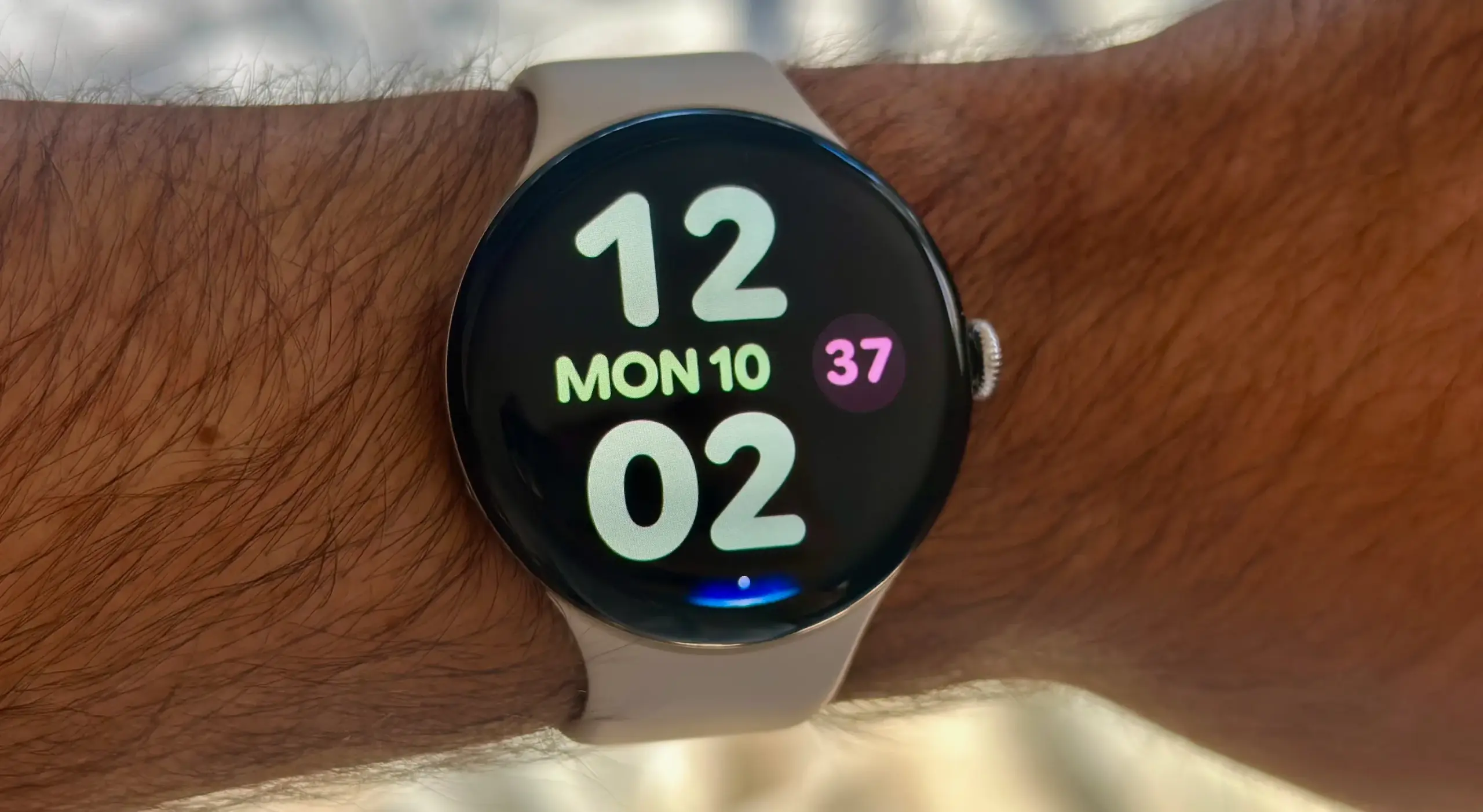 A photo of the Google Pixel Watch 4 on a man's wrist showing the Bold Utility watch face with large, colorful clock numbers and day/date info. At the display bottom is a blue Gemini Raise to Talk indicator.