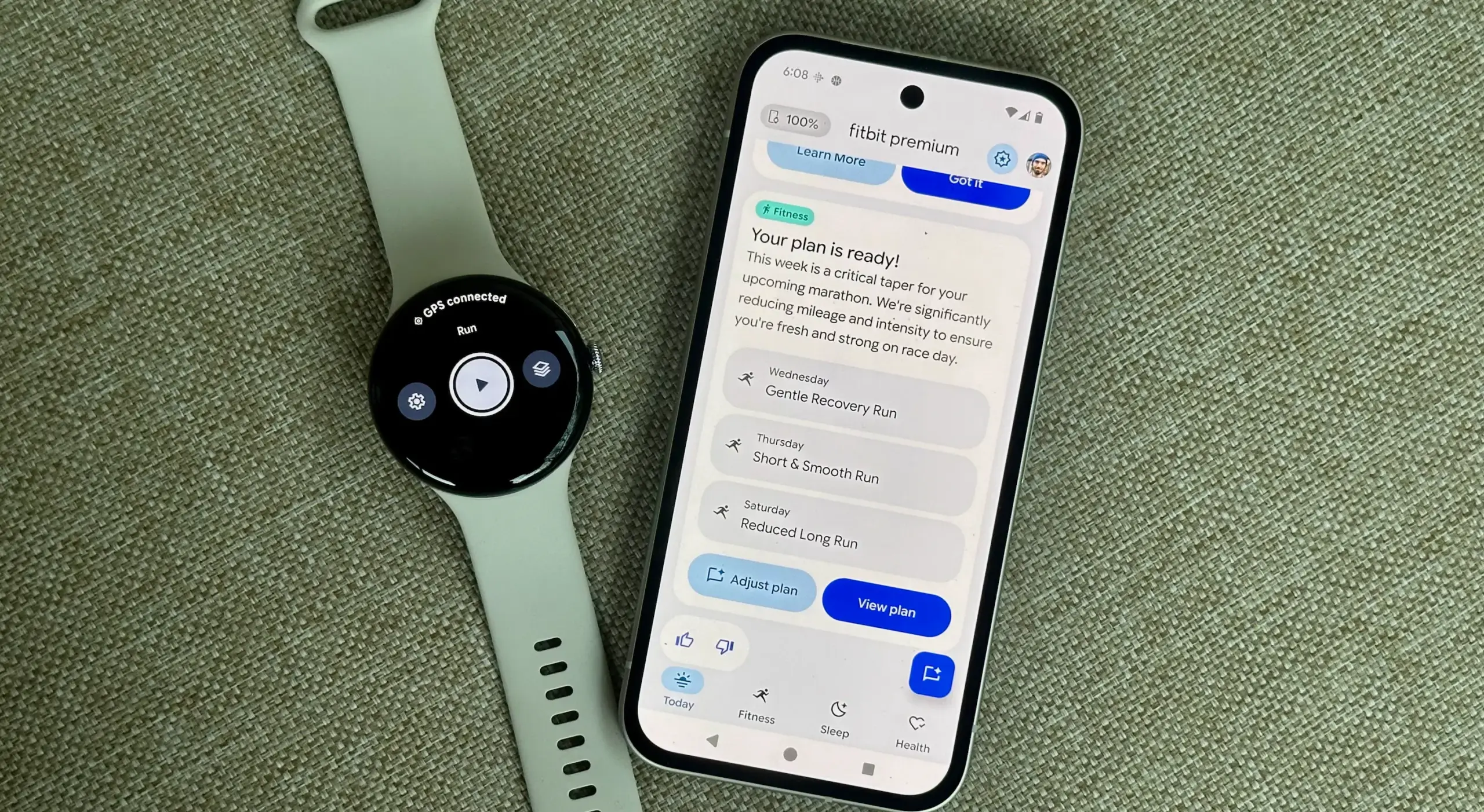 Google Pixel Watch 4 di samping Pixel 9a yang terbuka ke aplikasi Fitbit, menunjukkan preview Fitbit Premium Health Coach dengan tab Fitness terbuka.