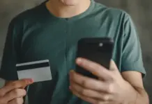 Pria muda memegang kartu kredit di satu tangan dan ponsel di tangan lainnya, simbol pengeluaran langganan aplikasi.