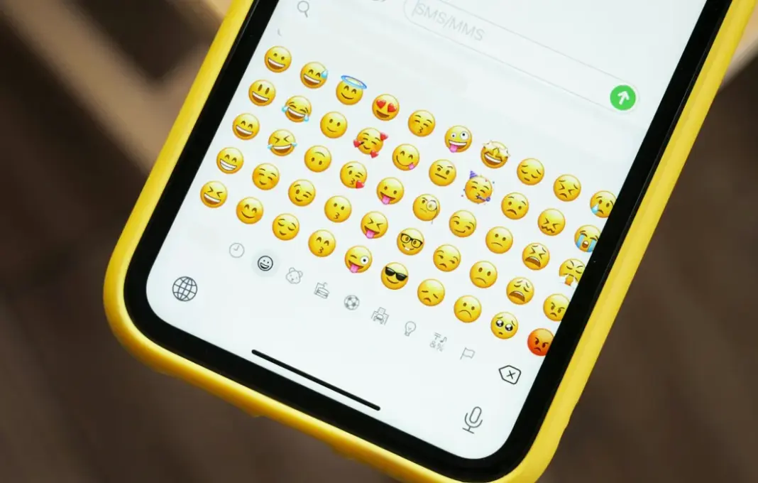 Ilustrasi beragam emoji populer 2025 melayang di layar ponsel, termasuk wajah dengan kantung mata dan hati merah.